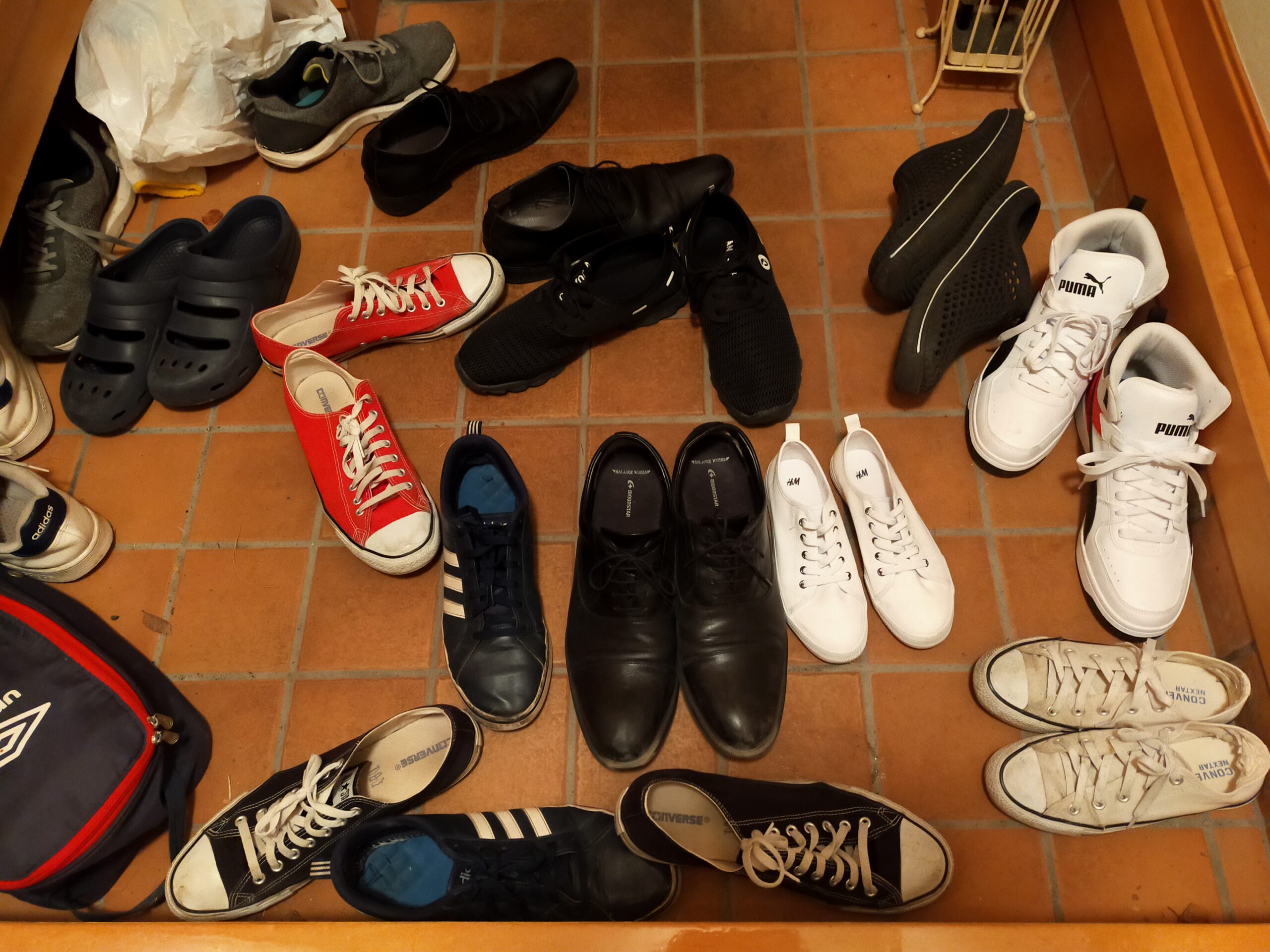 「玄関のタイル床いっぱいに、スニーカーやサンダル、革靴など多数の靴がランダムに広がっている写真。赤・黒・白・ネイビーなど色とりどりの靴が混ざり、複数人が出入りしたあとのにぎやかな玄関の様子が写っている。」