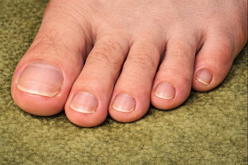 リアルな素足の指先を横からズームしたクローズアップ。5本すべての足指が並び、親指から小指までの爪の形や大きさの違いがはっきり分かる。特に小指の爪は他と比べて存在感があり、緑色のカーペットの上に自然な質感で描かれている。